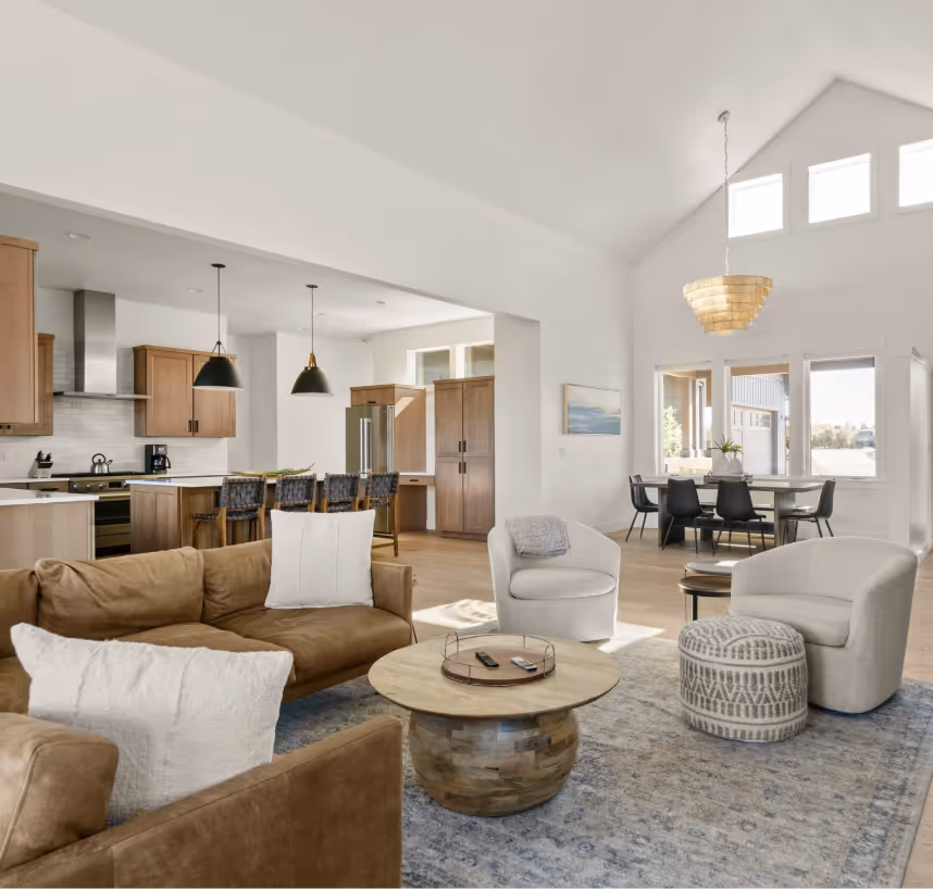 Open-plan living space with brown leather sofa, two white armchairs, patterned ottoman, round wooden coffee table, modern kitchen with island and bar stools, and dining area under a modern light fixture.