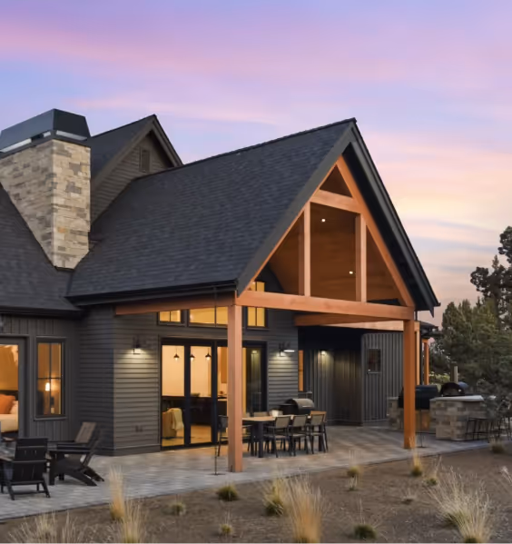 Modern house with dark siding, large windows, a wooden porch structure, and outdoor dining area at sunset.