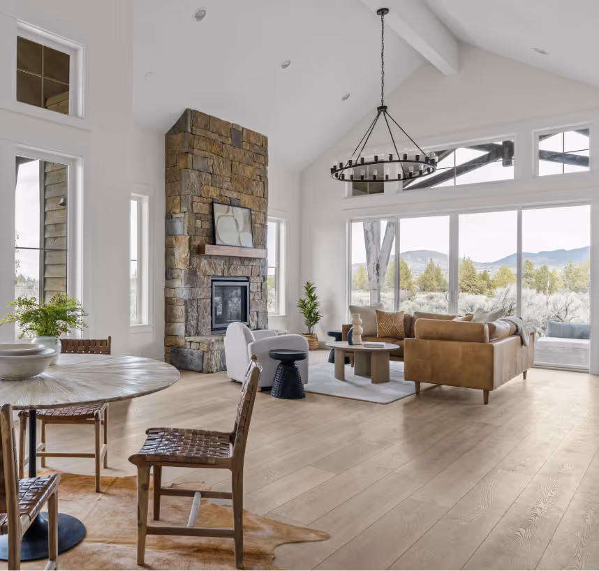 Bright living room with large windows, stone fireplace, leather sofa, and wooden dining table with woven chairs.