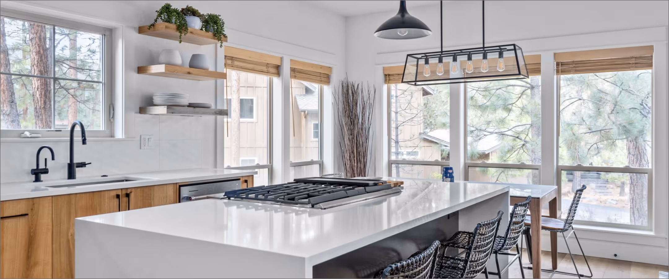Modern kitchen with a large white island featuring a built-in stovetop, black pendant lights, wooden cabinets, and large windows showing trees outside.