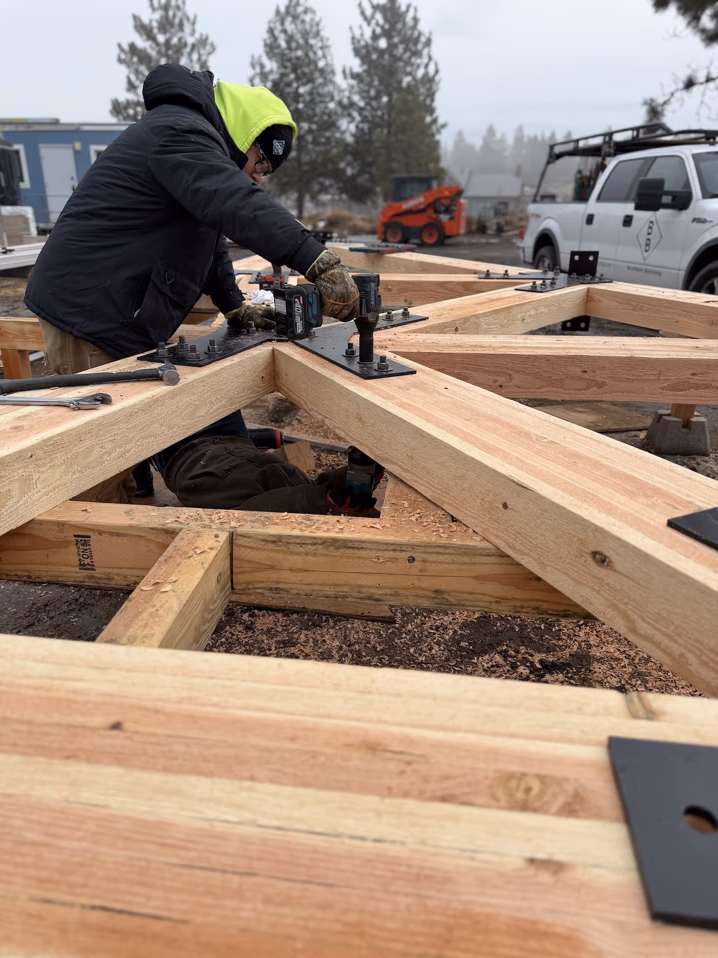 Construction workers assembling wooden beams using power drills at an outdoor site with a white truck and trees in the background.