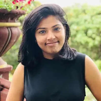 Young woman with shoulder-length black hair wearing a sleeveless black top, smiling outdoors with greenery and flowers in the background.