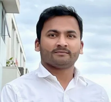 Man with short black hair and a trimmed beard wearing a white collared shirt against a cloudy sky background.
