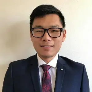 Smiling man wearing black glasses, a dark suit, white shirt, and patterned tie, standing against a plain light background.