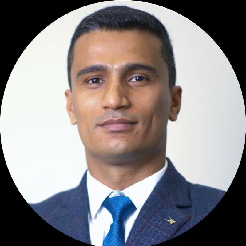 Portrait of a man with short black hair wearing a blue suit jacket, white shirt, and blue tie against a light background.