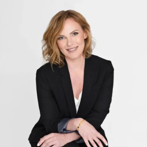 Smiling woman with shoulder-length light brown hair wearing a black blazer and white top, seated with hands crossed.