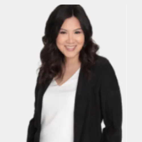 Smiling woman with long dark hair wearing a white top and black blazer against a light background.