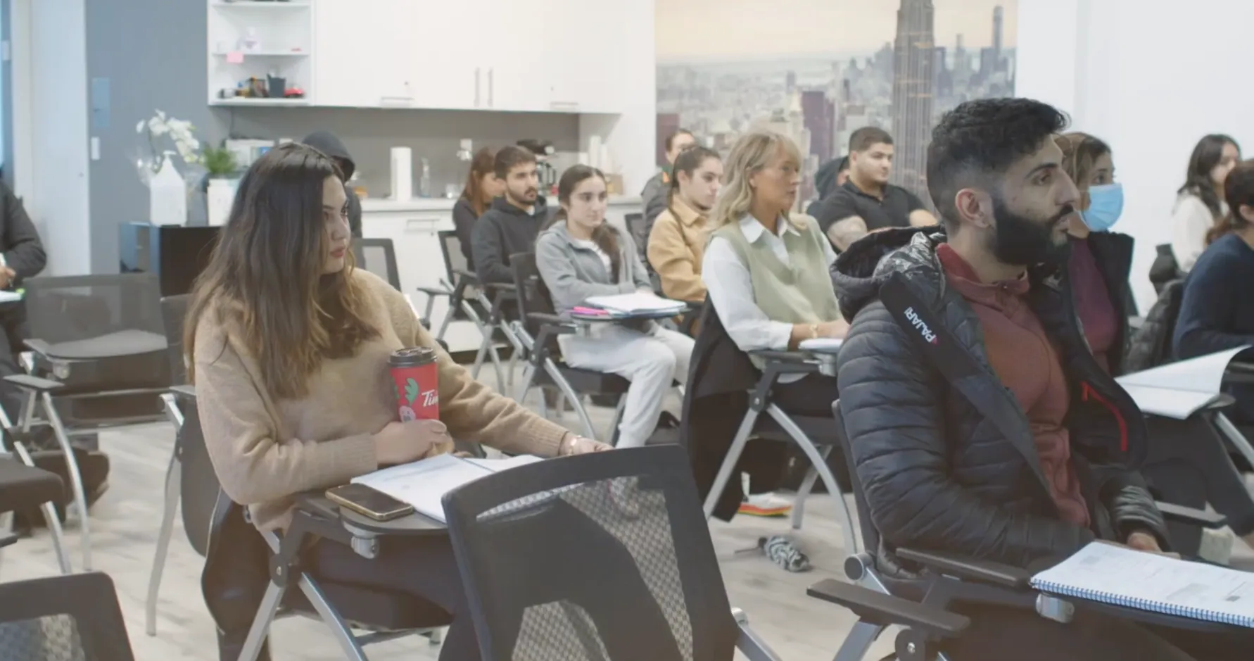 A diverse group of students attentively listening in a modern classroom with notebooks and a Tim Hortons cup visible.