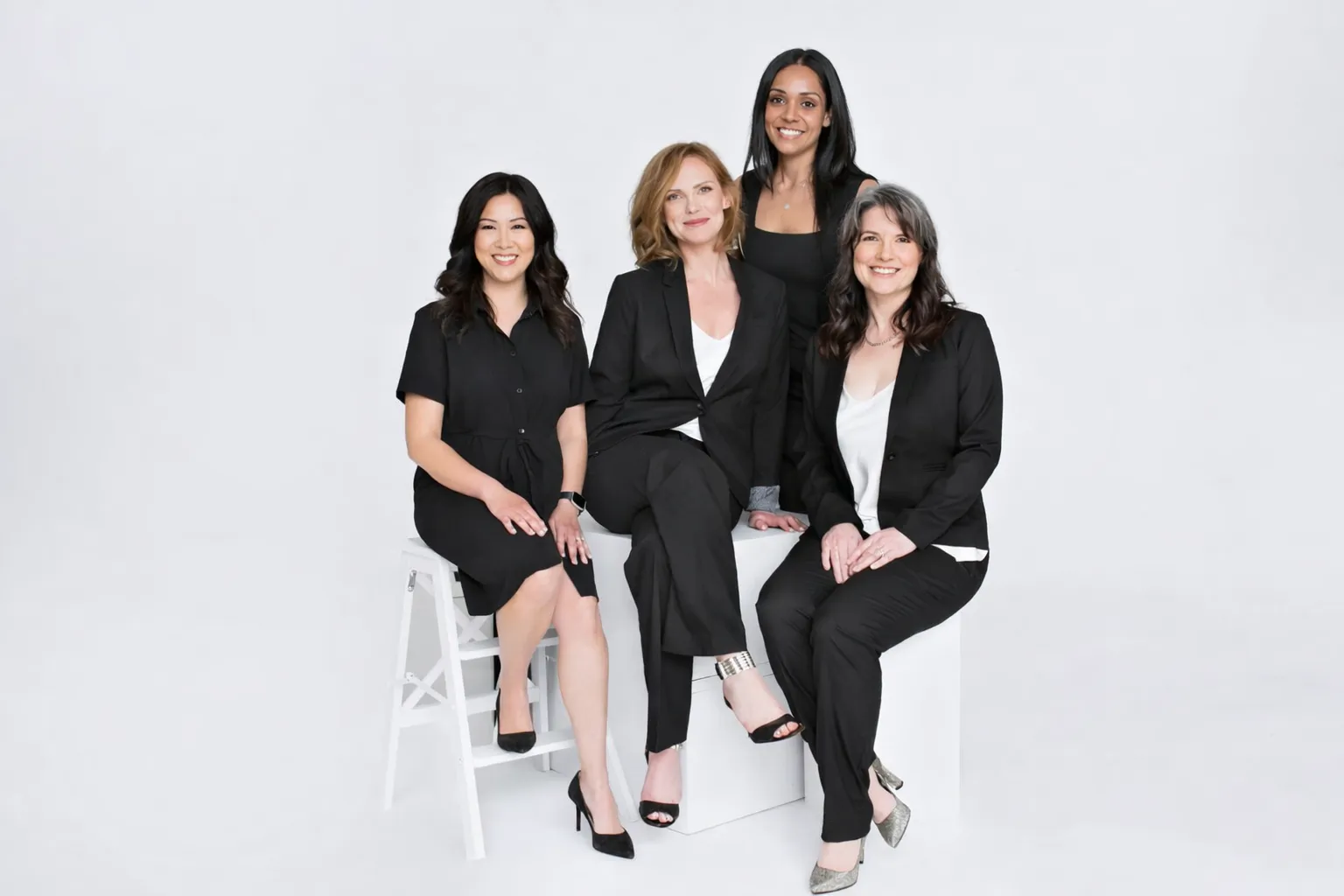 Four women dressed in black business attire posing against a light gray background, two seated on white props and two standing behind.