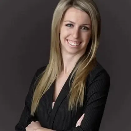 Smiling woman with long blonde hair wearing a black blazer, standing with arms crossed against a dark background.