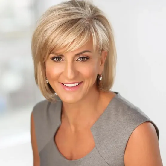 Smiling woman with short blonde hair wearing a gray sleeveless top in a bright indoor setting.