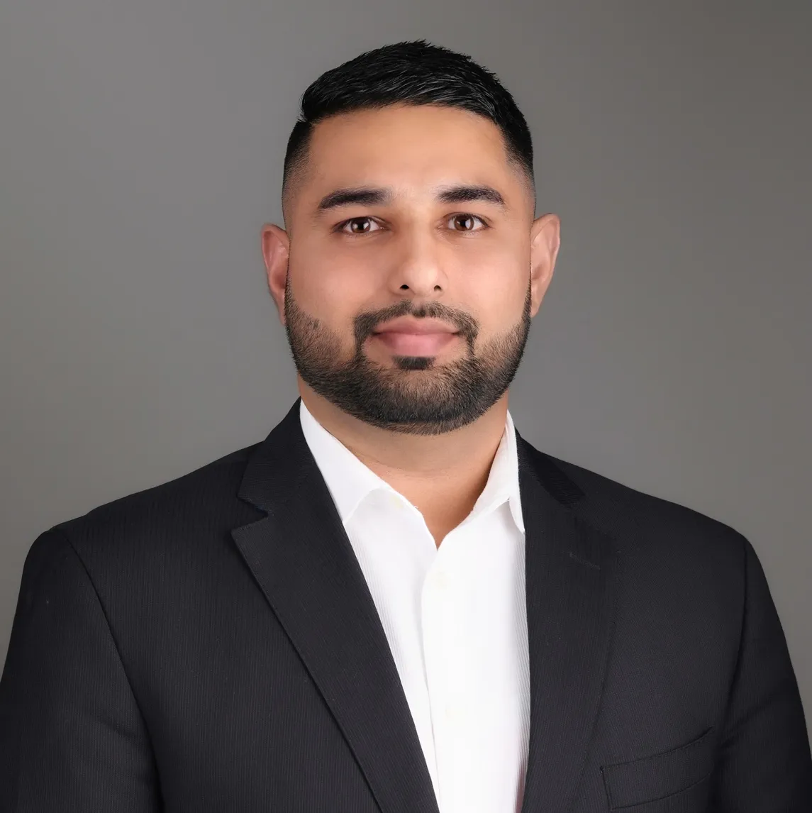 Portrait of a man with short black hair and beard wearing a black suit jacket and white shirt against a gray background.