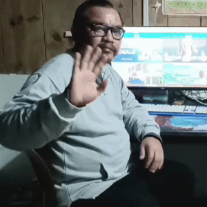 Man wearing glasses and a light gray sweater waving at the camera while sitting in front of a computer screen.
