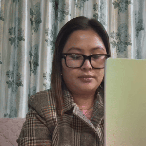 Woman with glasses and straight hair wearing a plaid jacket, looking down at a laptop in front of a floral curtain.