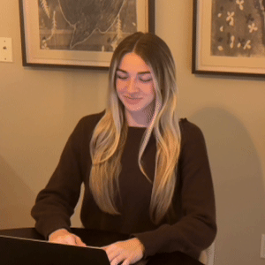 Woman with long blonde hair wearing a dark sweater, smiling and typing on a laptop indoors.
