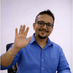 Man with glasses and beard waving his hand while smiling, wearing a blue shirt.