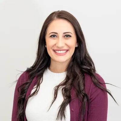 Smiling woman with long dark hair wearing a white top and maroon cardigan against a light gray background.