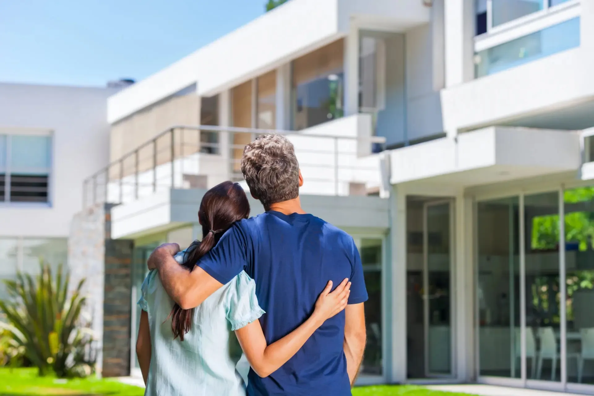 Couple embracing and looking at a modern house with large windows and a balcony.