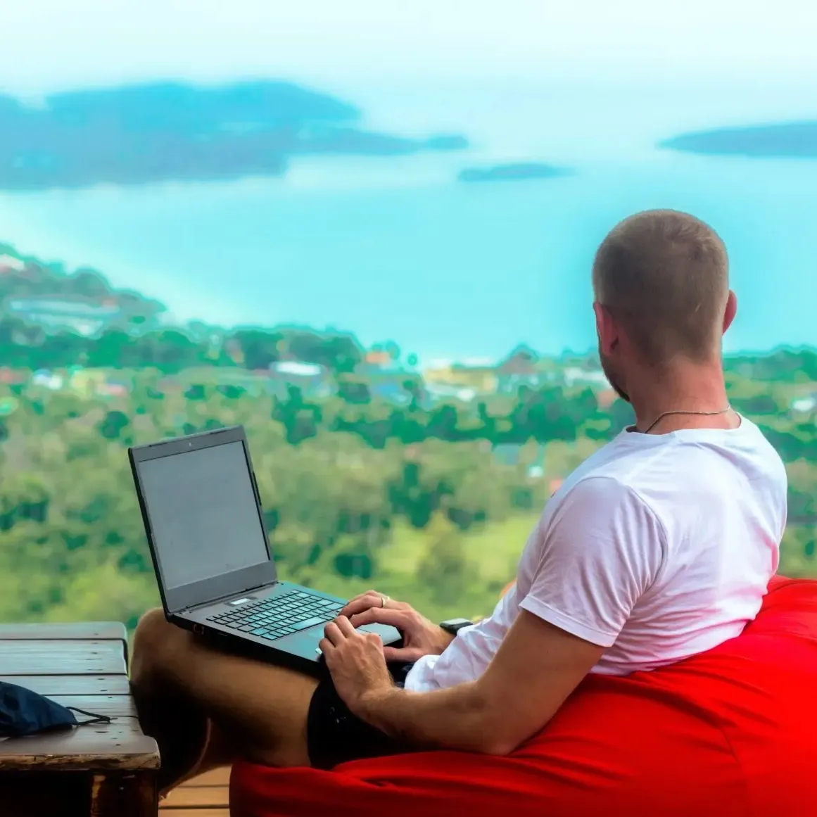 Man in white t-shirt sitting on a red bean bag using a laptop with a blurred scenic view of water and greenery in the background.
