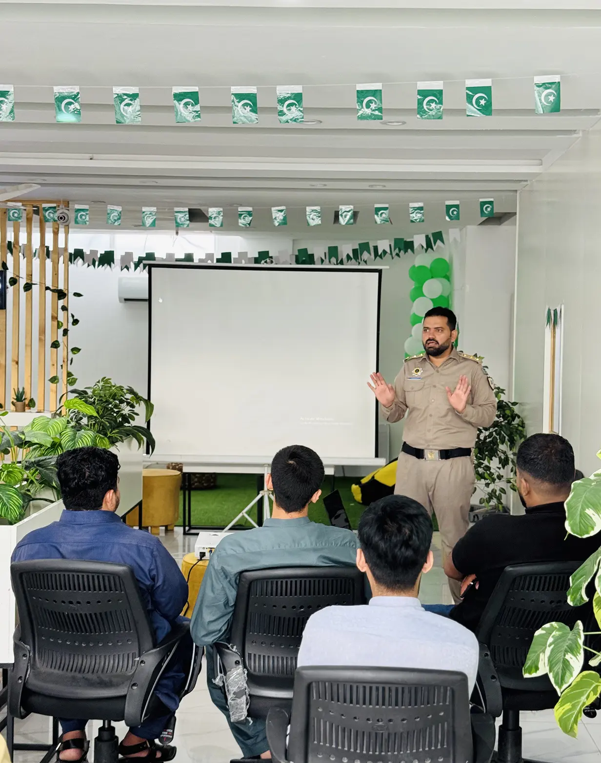 A man in uniform giving a presentation to an audience of four seated men in an indoor room decorated with green and white flags and balloons.