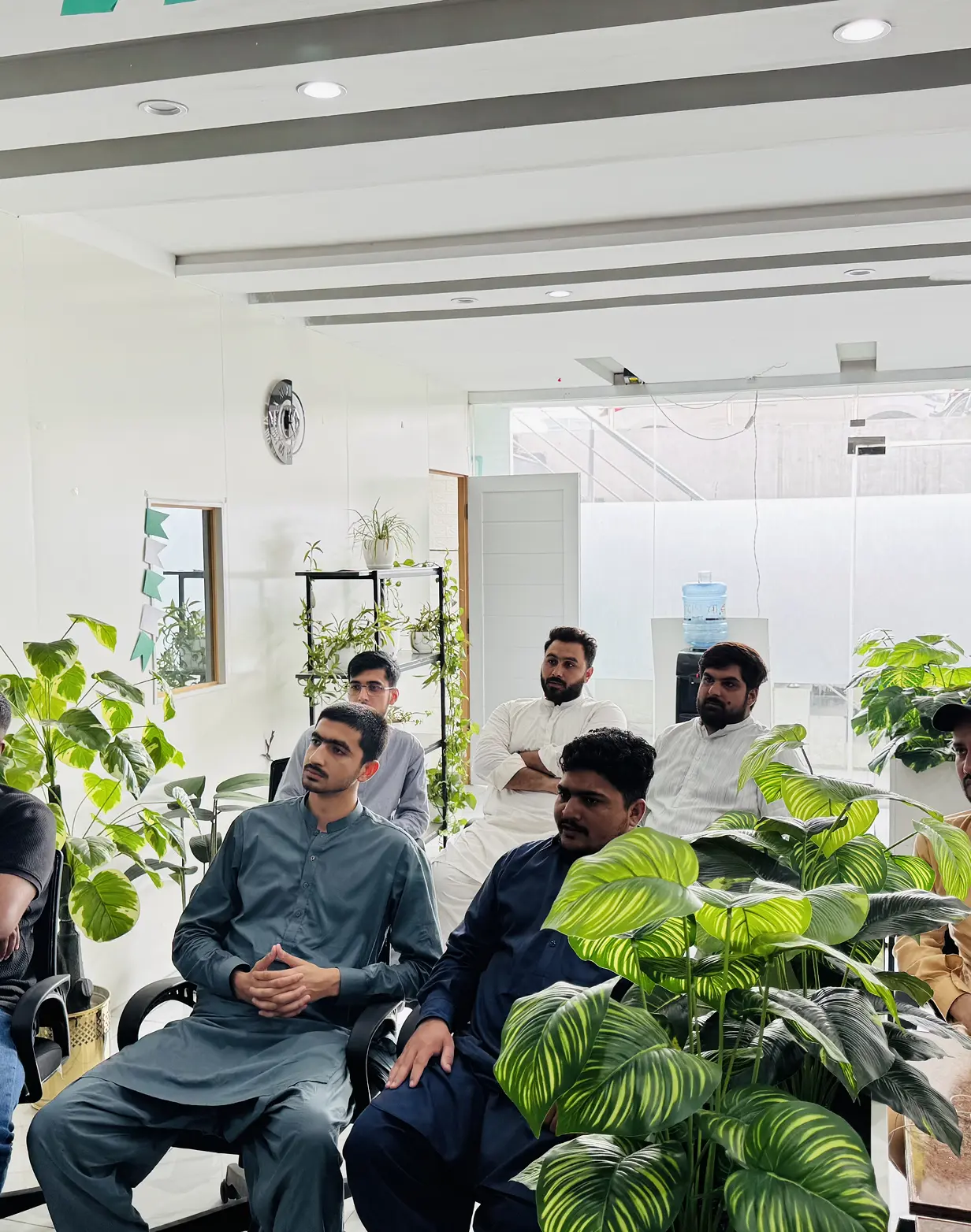 Group of men sitting attentively in a bright room decorated with green potted plants and white walls.