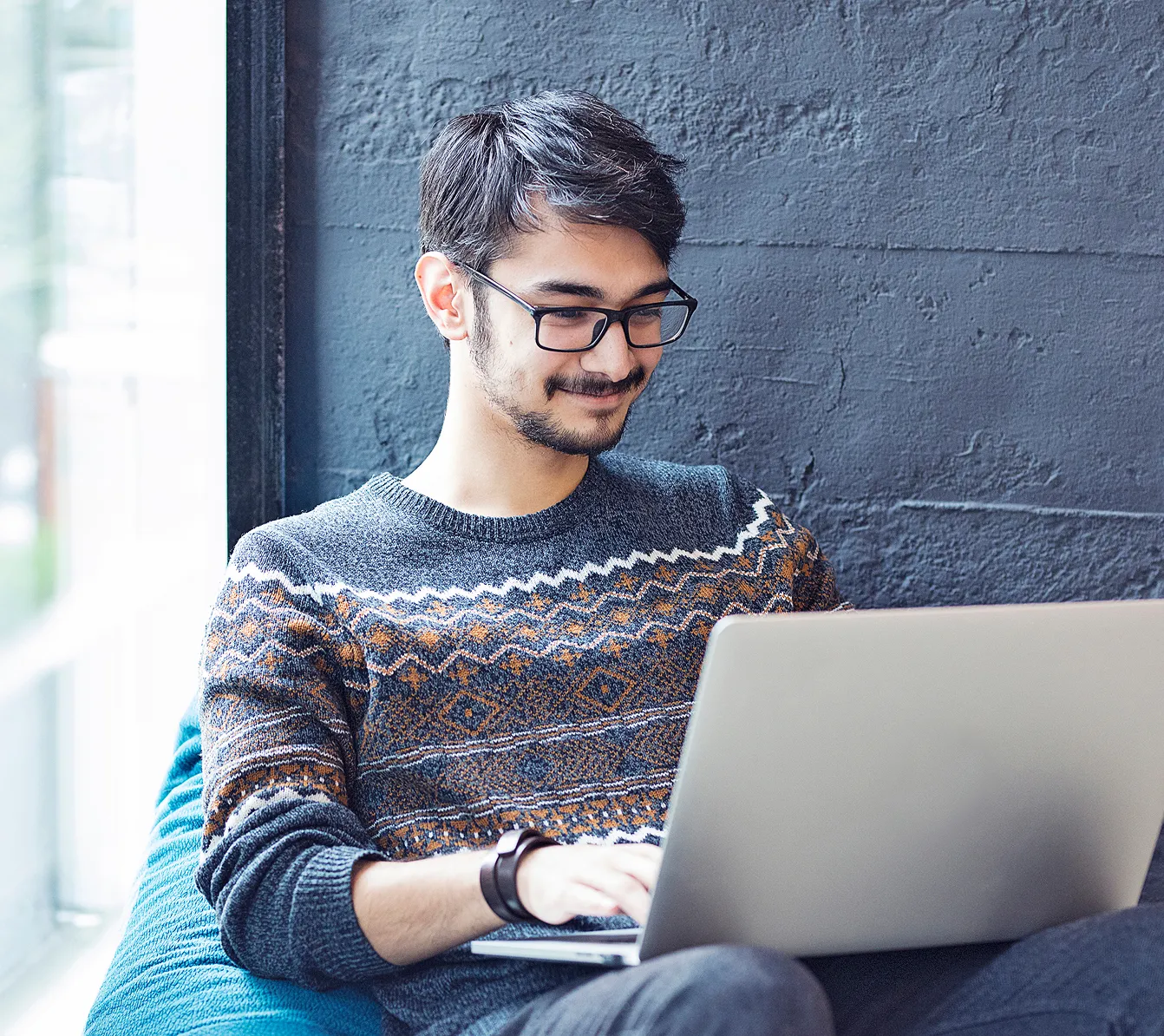 Man wearing glasses and a patterned sweater working on a laptop while sitting near a window.