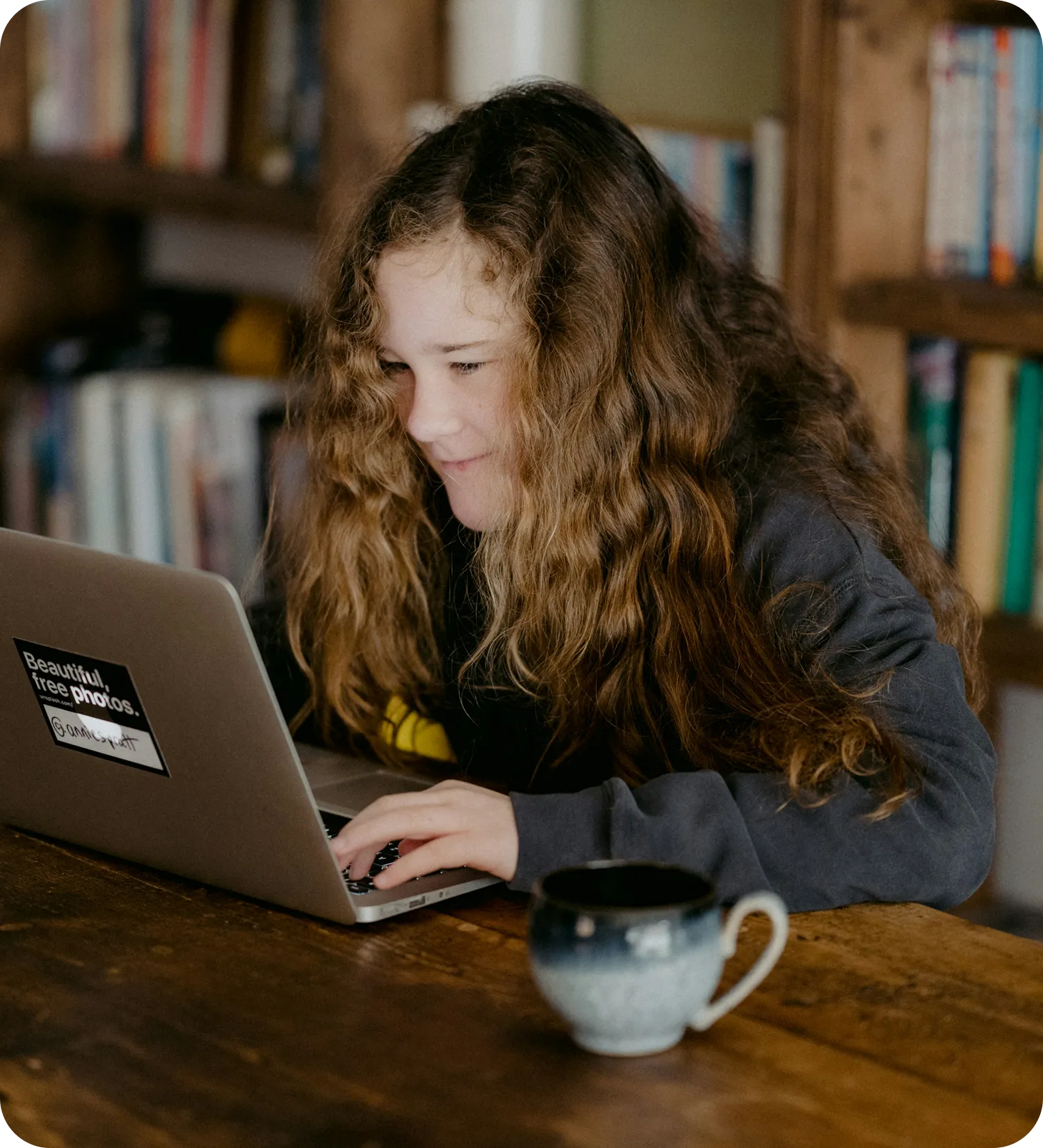 Young person with long curly hair using a laptop at a wooden table with a ceramic mug nearby.