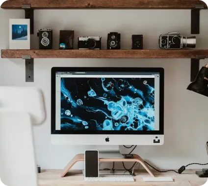 Desk workspace with a computer displaying abstract blue and black art and vintage cameras on wooden shelves above.