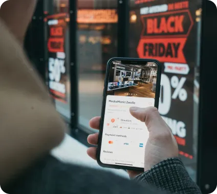 Person holding a smartphone displaying store details with Black Friday sale signs blurred in the background.