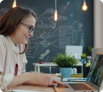 Young woman with glasses using a laptop at a desk with a small plant and hanging lights.