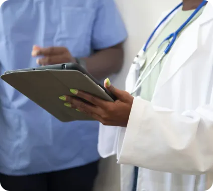 Healthcare professionals, one in a white coat with stethoscope holding a tablet, standing with another in blue scrubs.