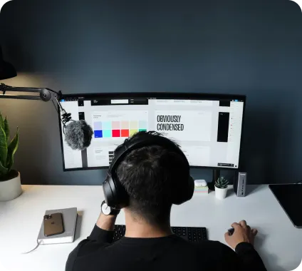Person wearing headphones and sitting at a desk working on a computer with a microphone attached to the monitor.