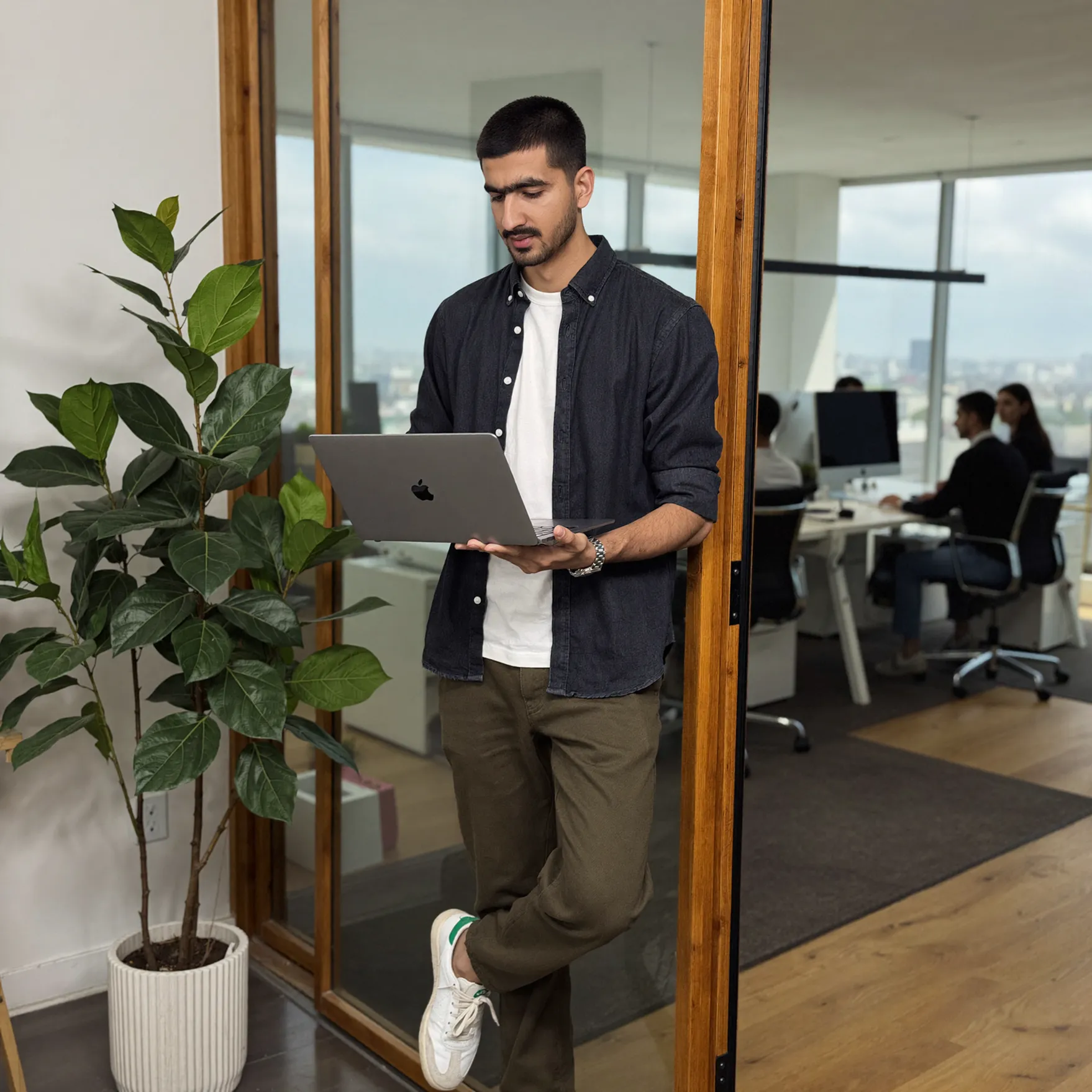 Man standing and working on a laptop near a plant in a modern office with coworkers inside.