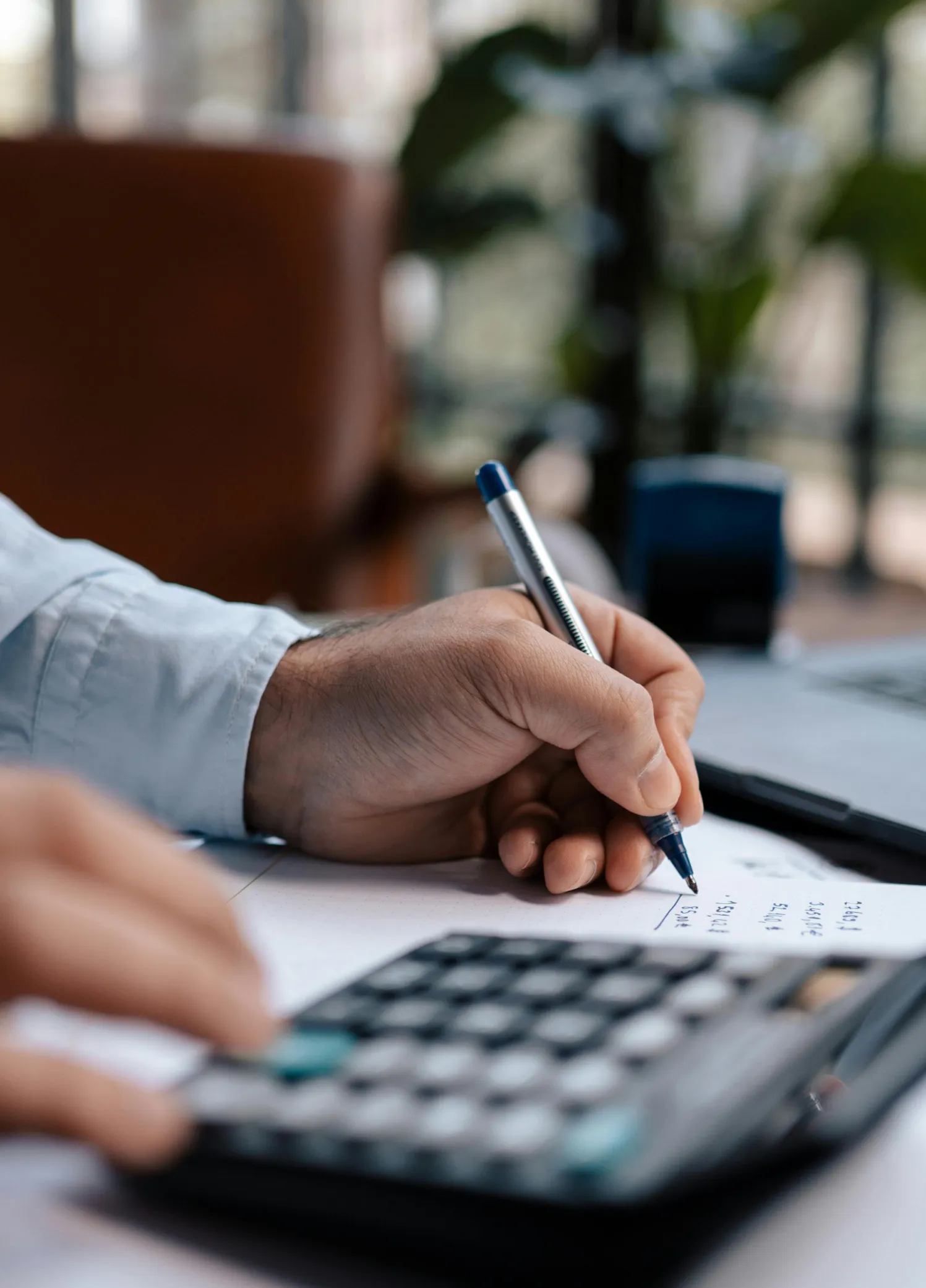 Person using a calculator and writing calculations on paper with a blue pen.
