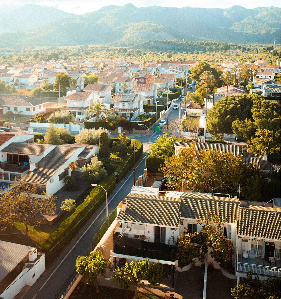 Suburban neighborhood with houses, trees, and mountains in the background under bright sunlight.