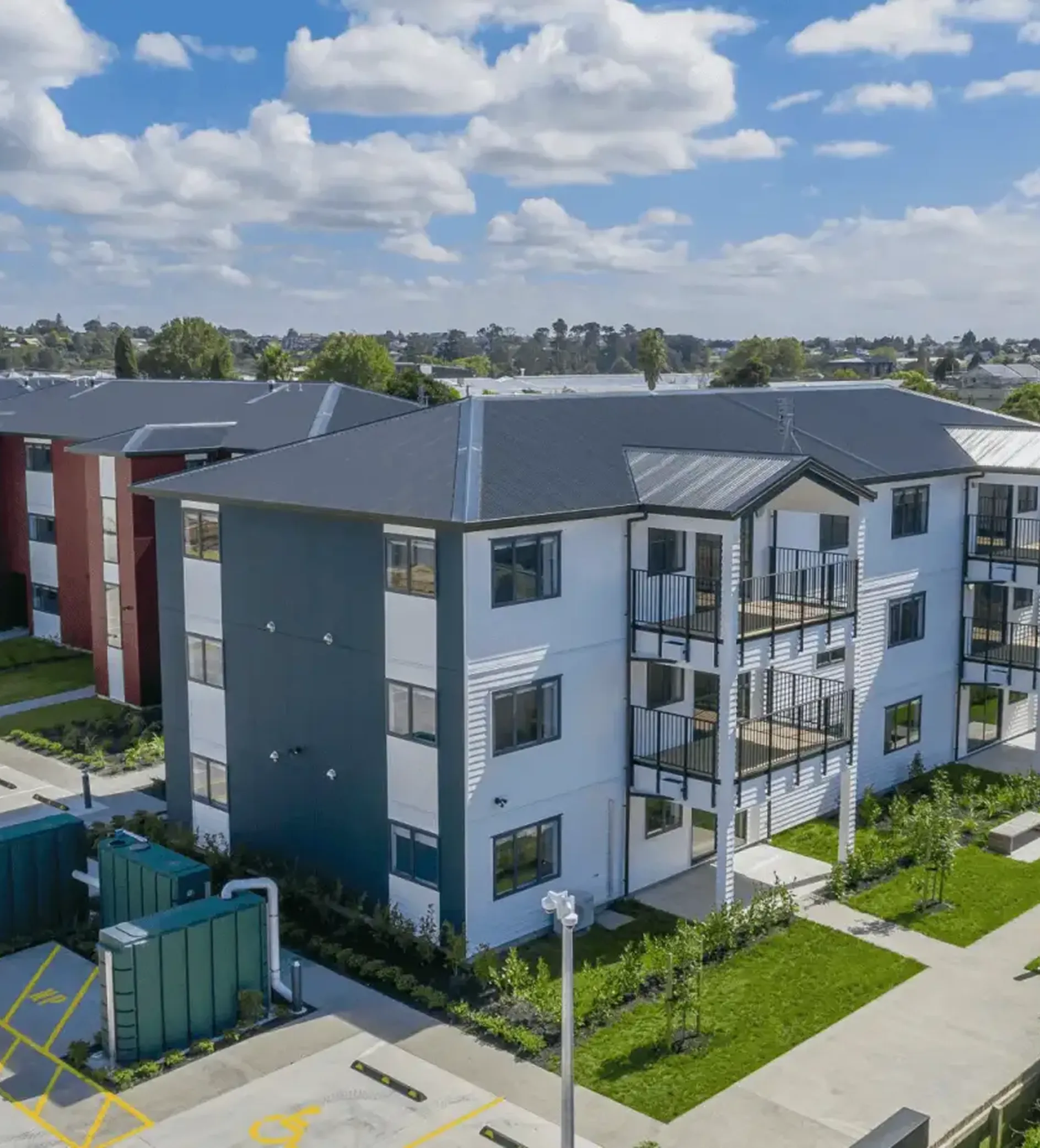 Modern three-story apartment building with balconies, parking area, and landscaped greenery.