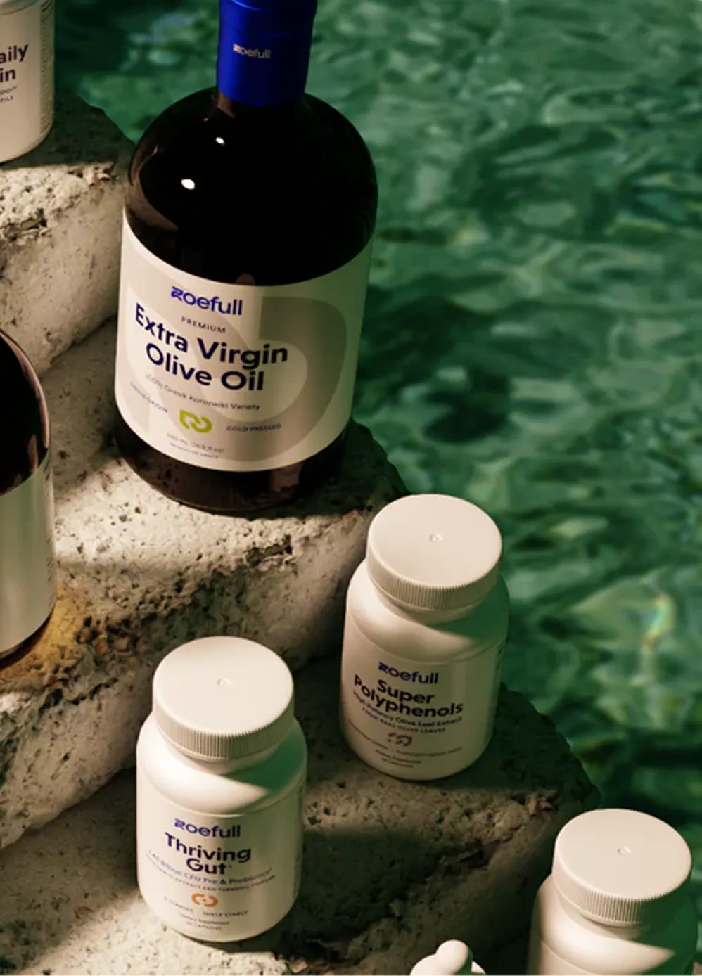 Bottles of Zoefull Extra Virgin Olive Oil and supplements on stone steps near green water.
