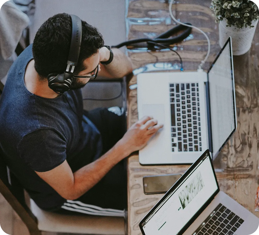 Man wearing headphones uses a laptop at a wooden table with another laptop and a smartphone nearby.