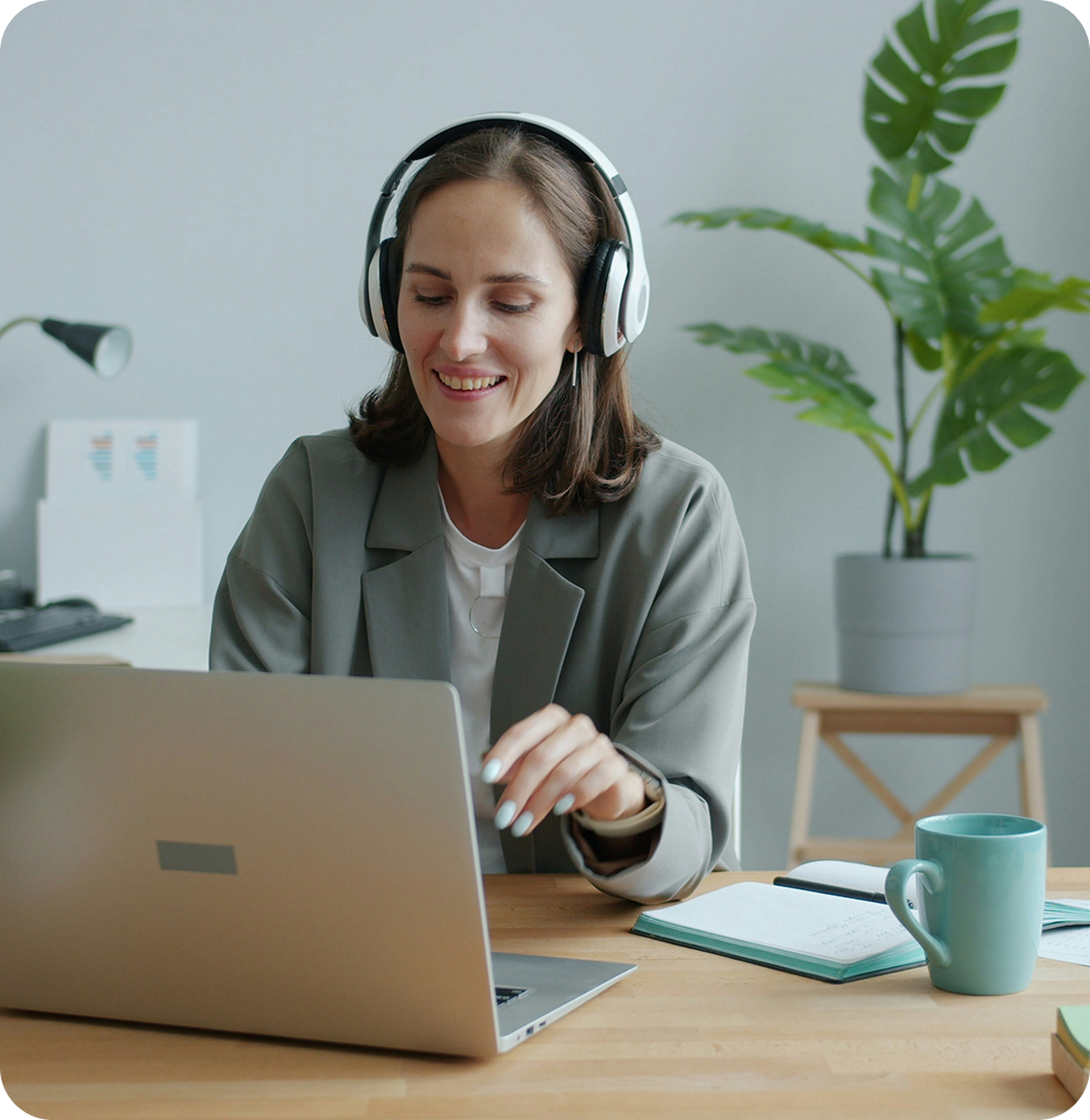 Smiling woman wearing headphones working on a laptop at a desk with a mug and notebook.