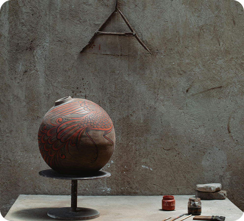 Round pottery vase with red bird design on rotating stand beside paint jars and brushes.