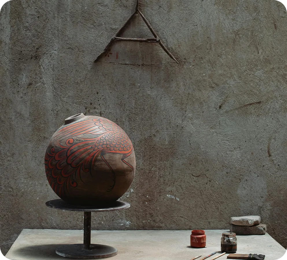 Round pottery vase with red bird design on rotating stand beside paint jars and brushes.