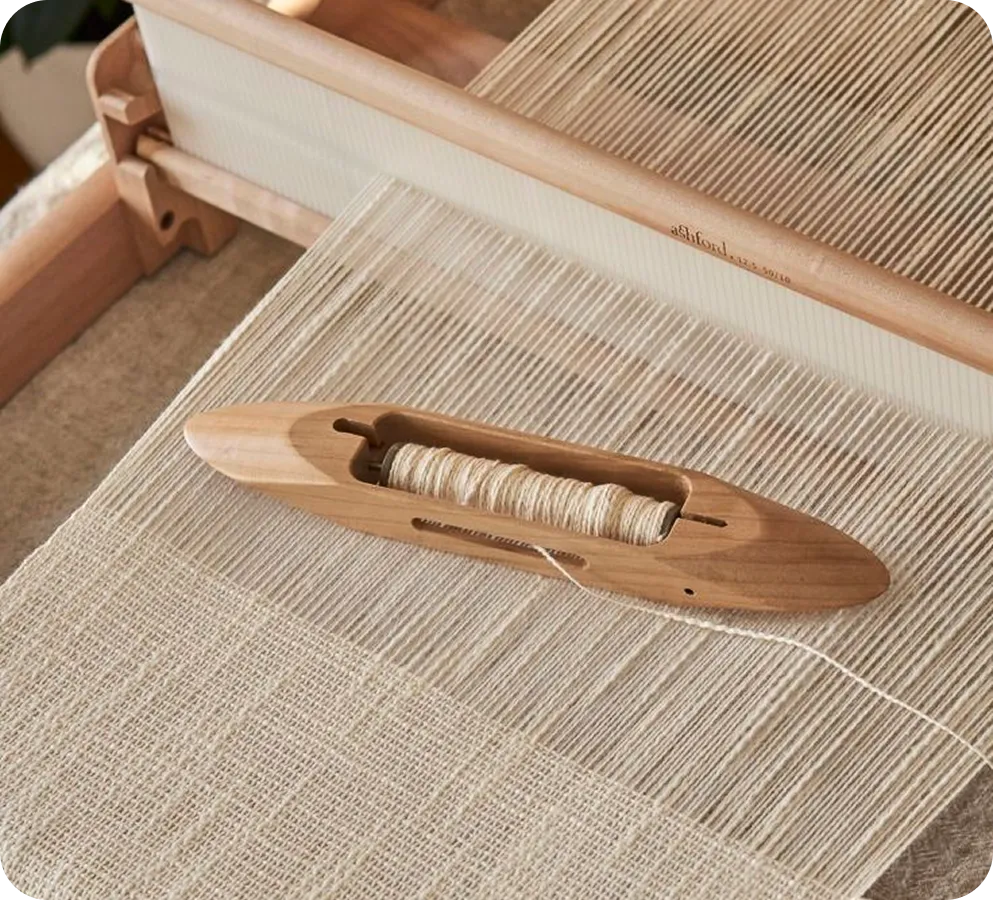 Wooden shuttle with thread on a loom weaving a beige textile fabric.