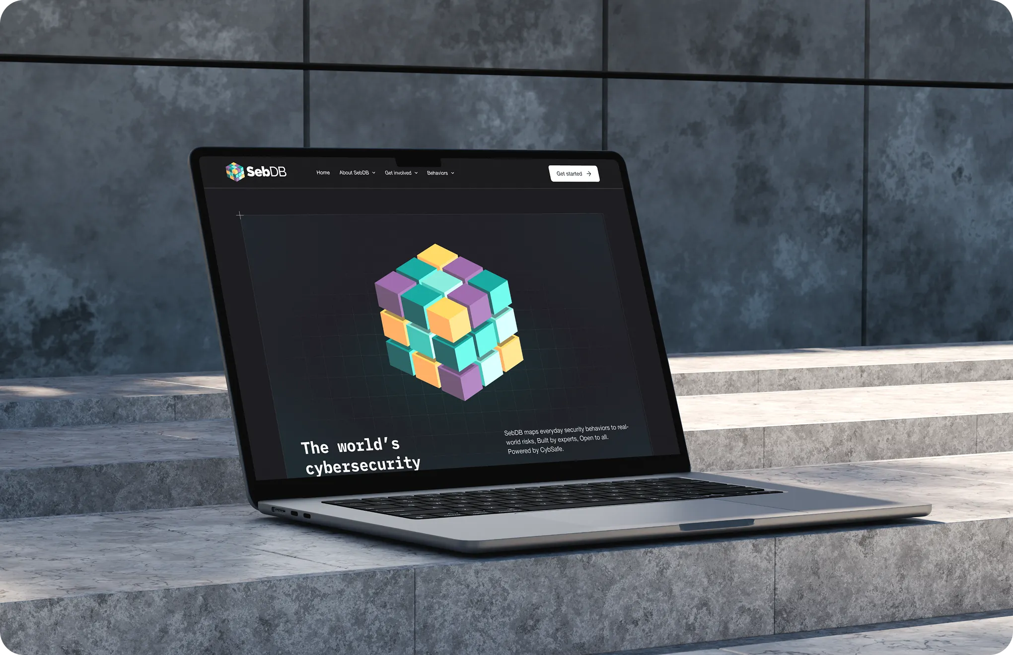 MacBook on stone steps displaying SebDB website with a colorful 3D cube graphic.