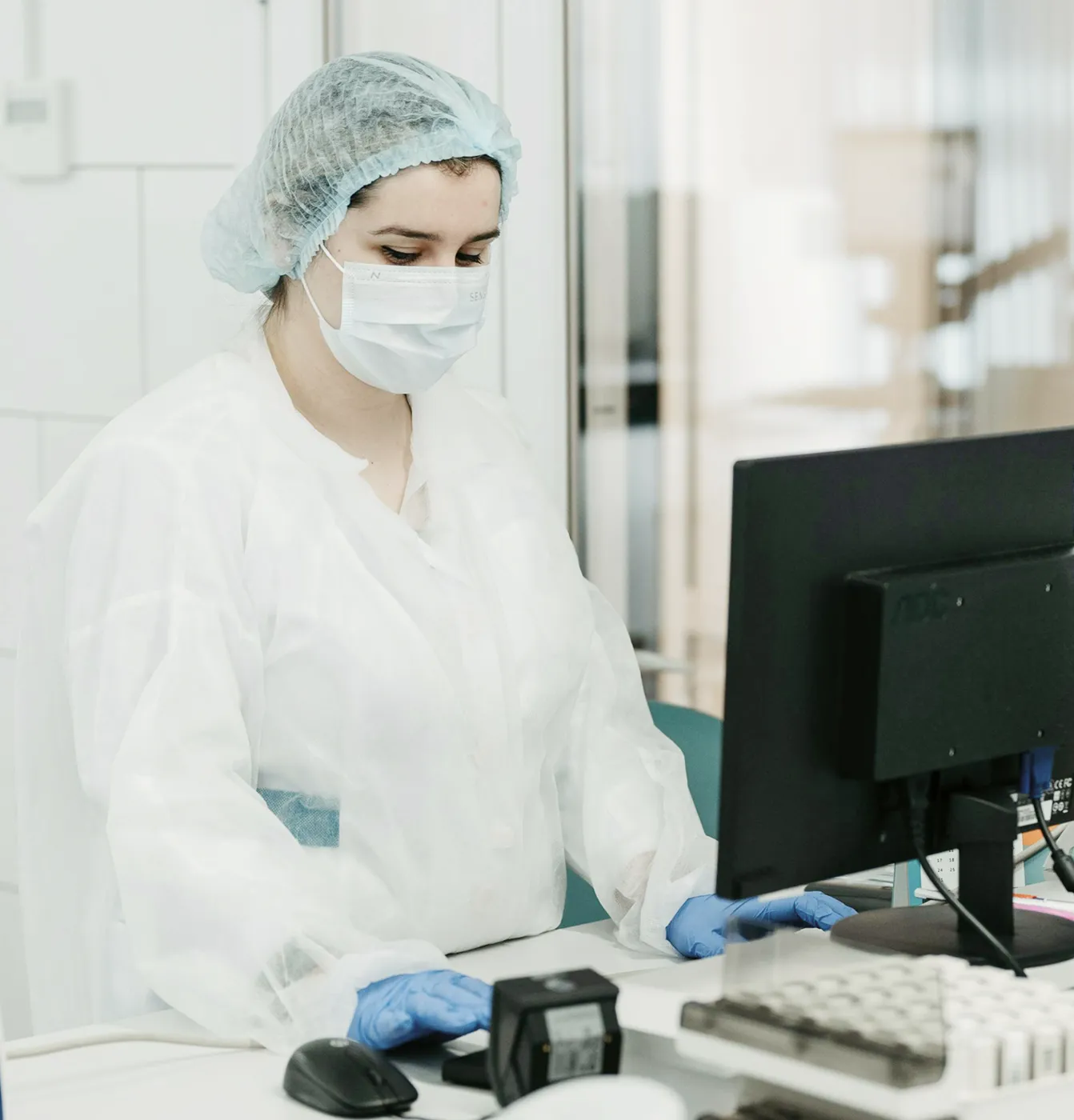 Medical worker in protective gown, mask, and hairnet working at a computer in a lab.