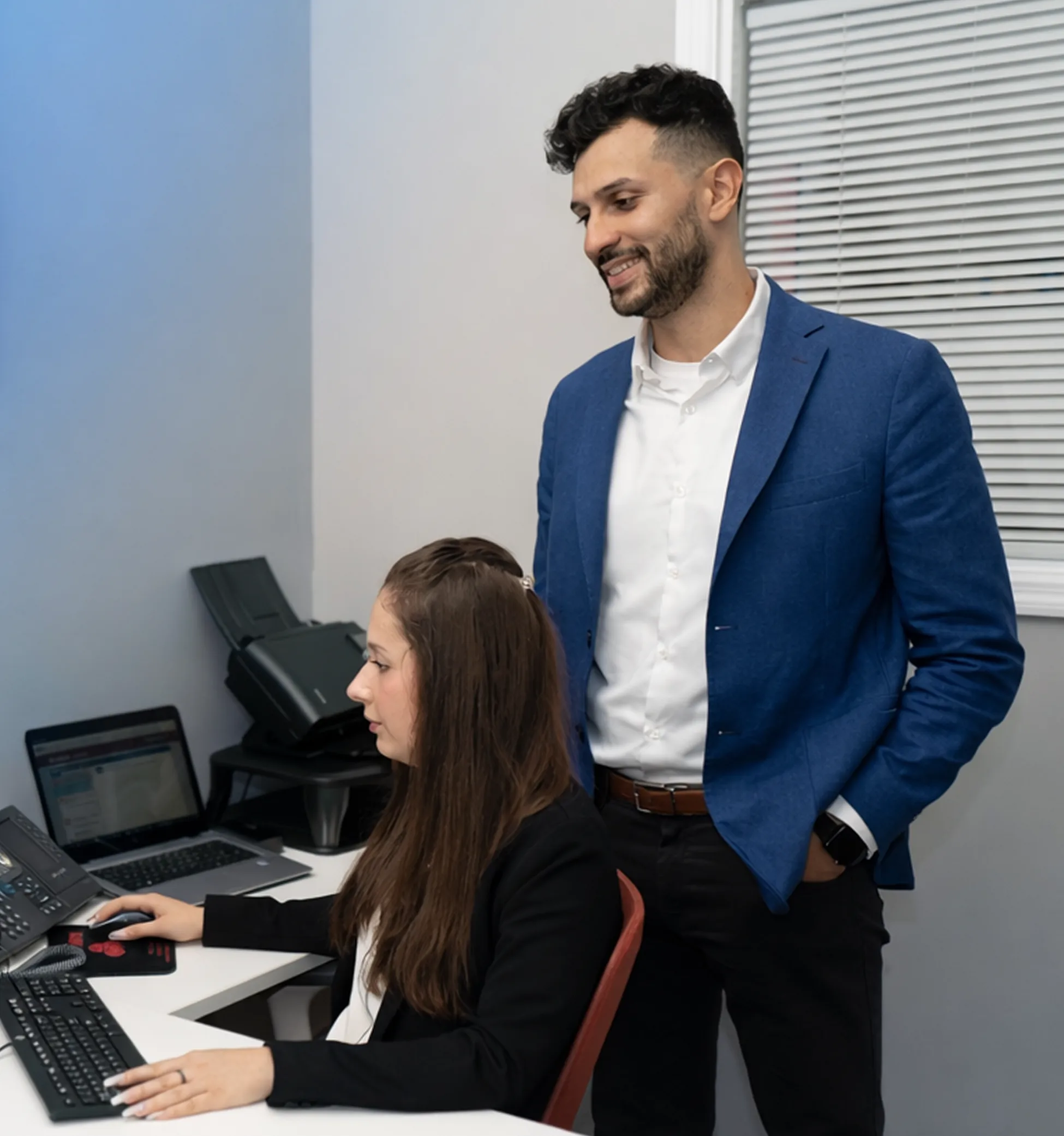 Woman using desktop computer while man stands beside her smiling in an office setting.
