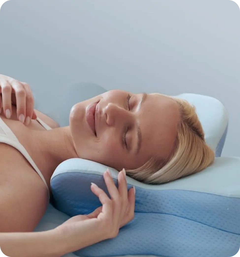Smiling woman lying down with eyes closed, resting her head on a blue contour memory foam pillow.