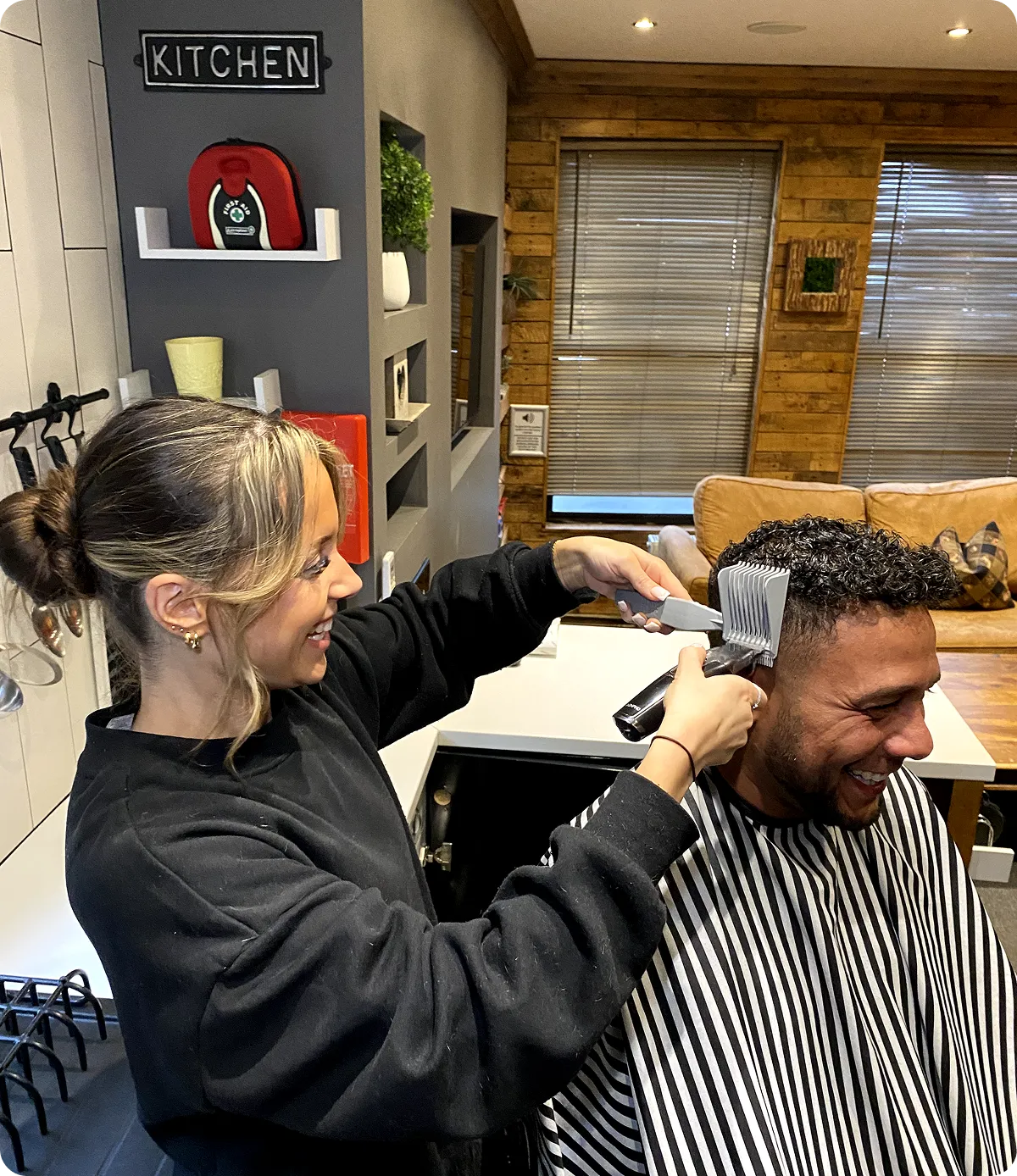 A woman cutting a man’s curly hair with clippers inside a cozy room with wood walls and kitchen sign.