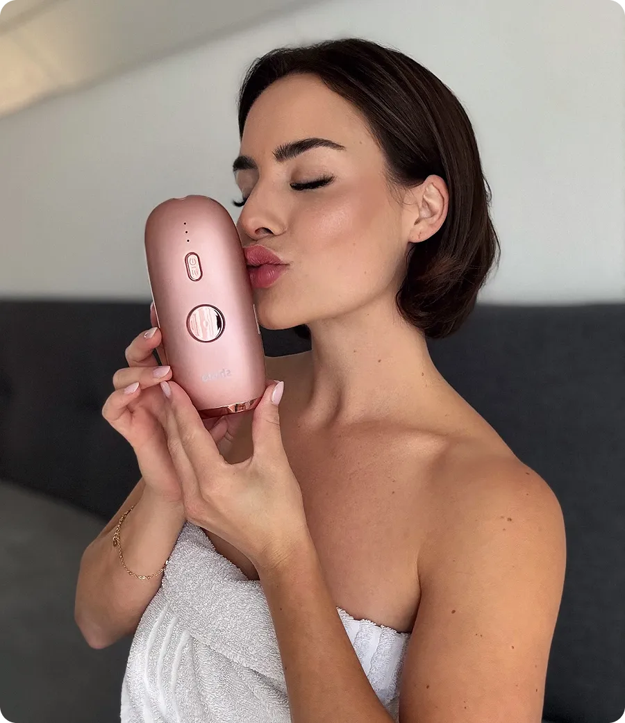 Woman wrapped in towel kissing a rose gold skincare or beauty device near her face.