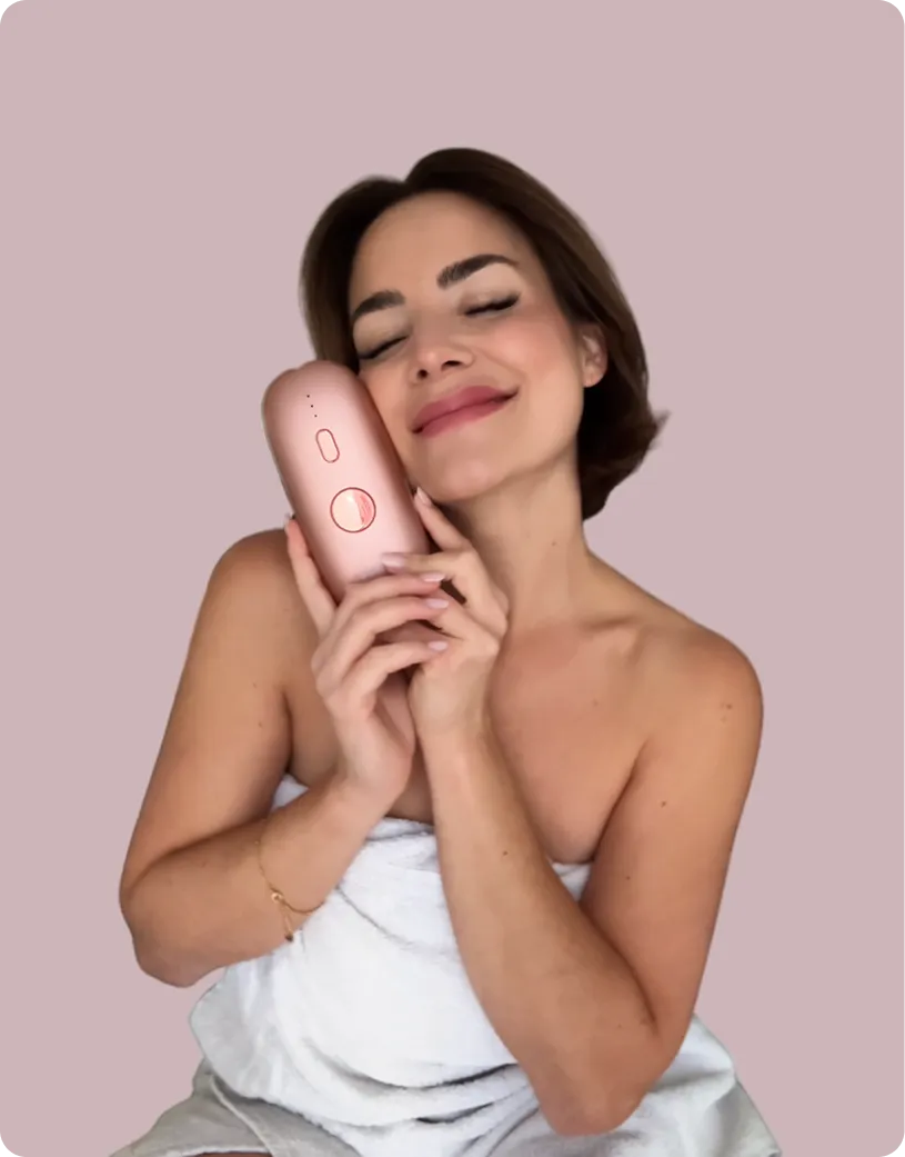 Smiling woman wrapped in a towel holding a rose gold device close to her face with eyes closed.