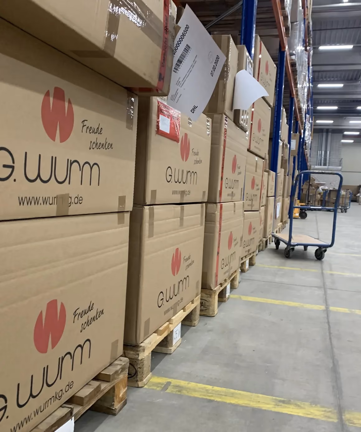 Warehouse aisles with stacks of G.wurm cardboard boxes on wooden pallets and a empty flat cart.
